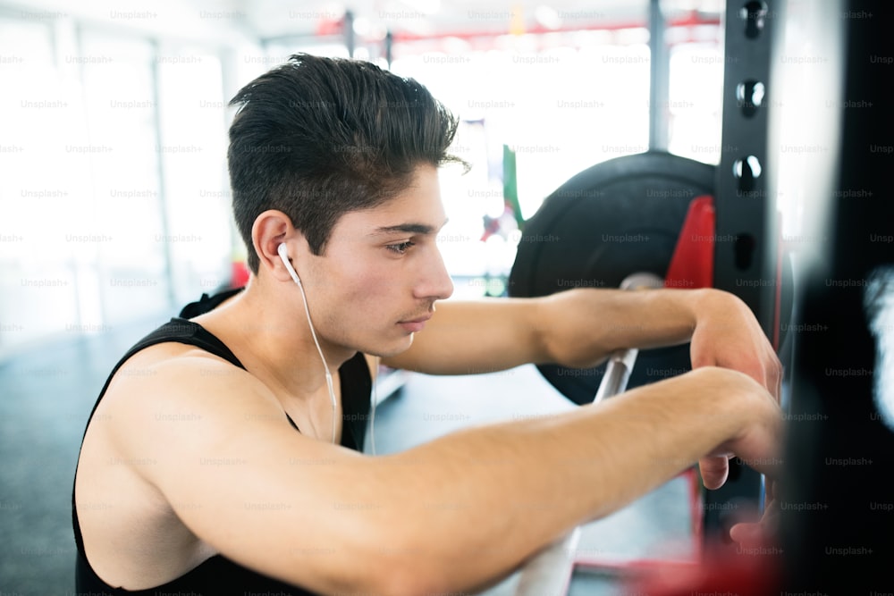 Hispanic fitness man in gym at the heavy barbell, resting, earphones in his ears, listening music