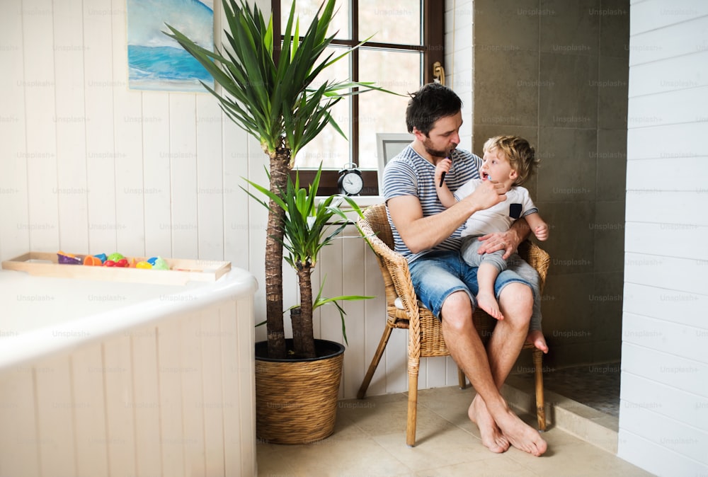 Father and a toddler boy sitting on a chair and brushing their teeth in the bathroom at home. Paternity leave.
