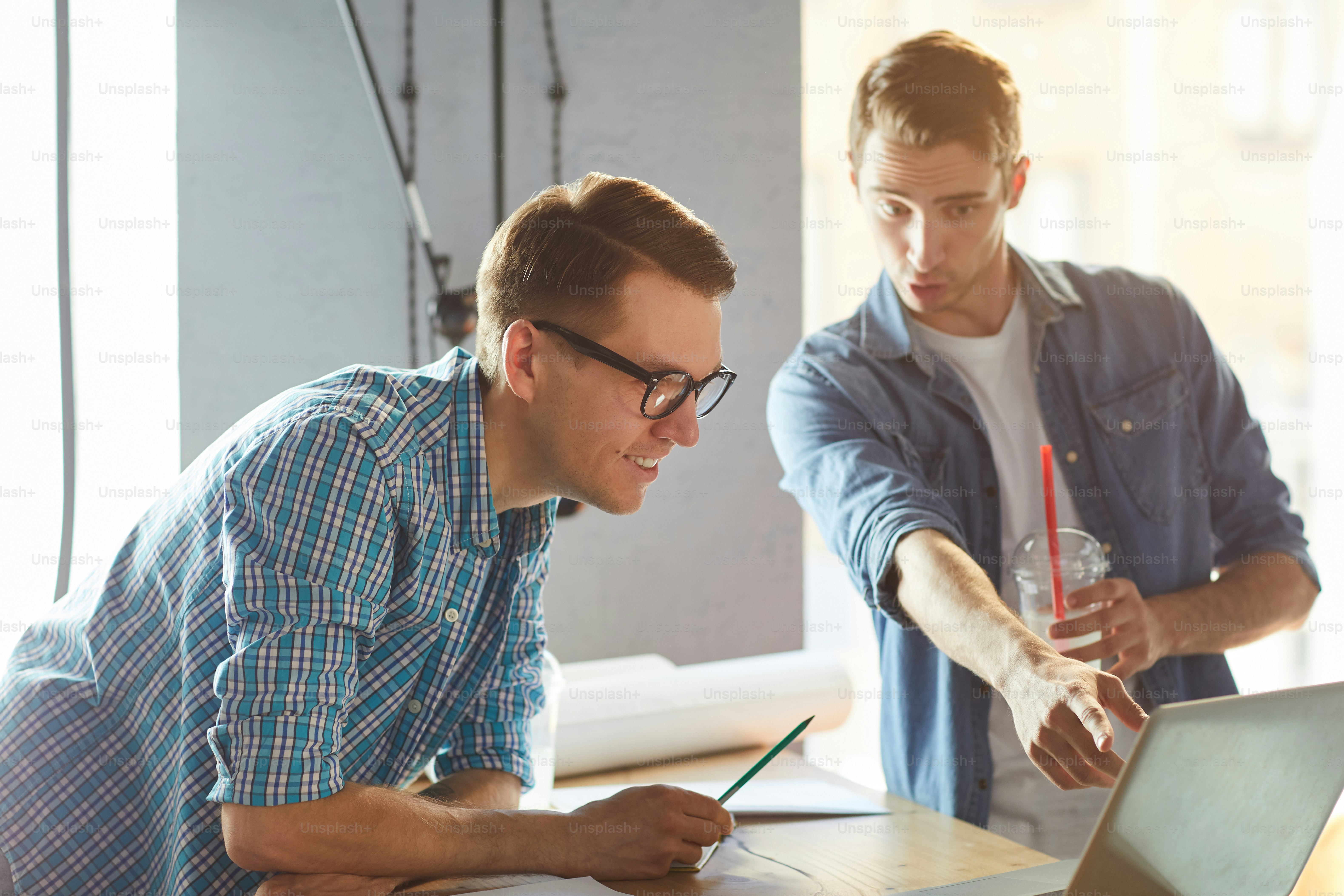 Young architect pointing at laptop display while explaining his colleague online data at meeting