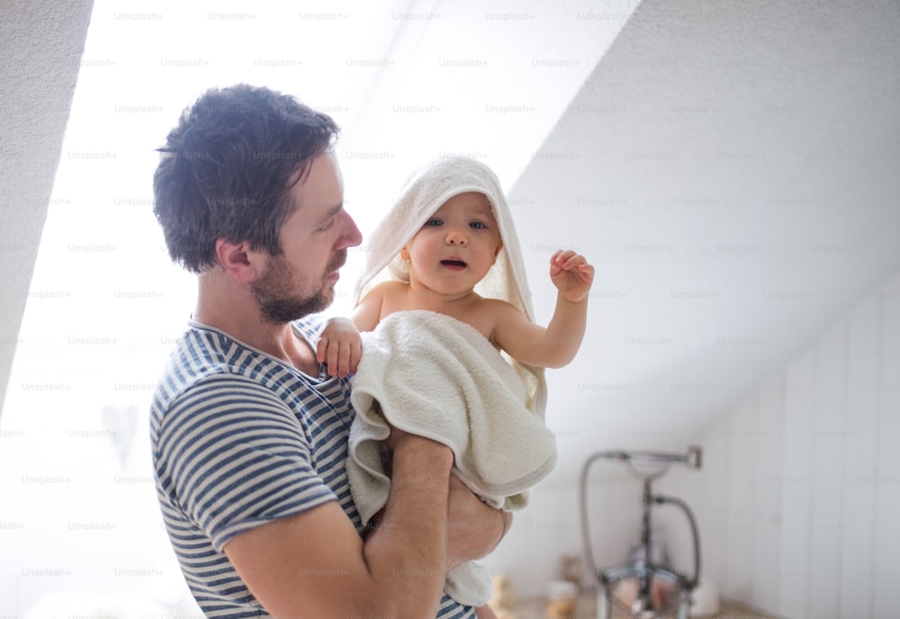 Vater mit einem glücklichen Kleinkind, eingewickelt in ein Handtuch in einem Badezimmer zu Hause. Vaterschaftsurlaub.
