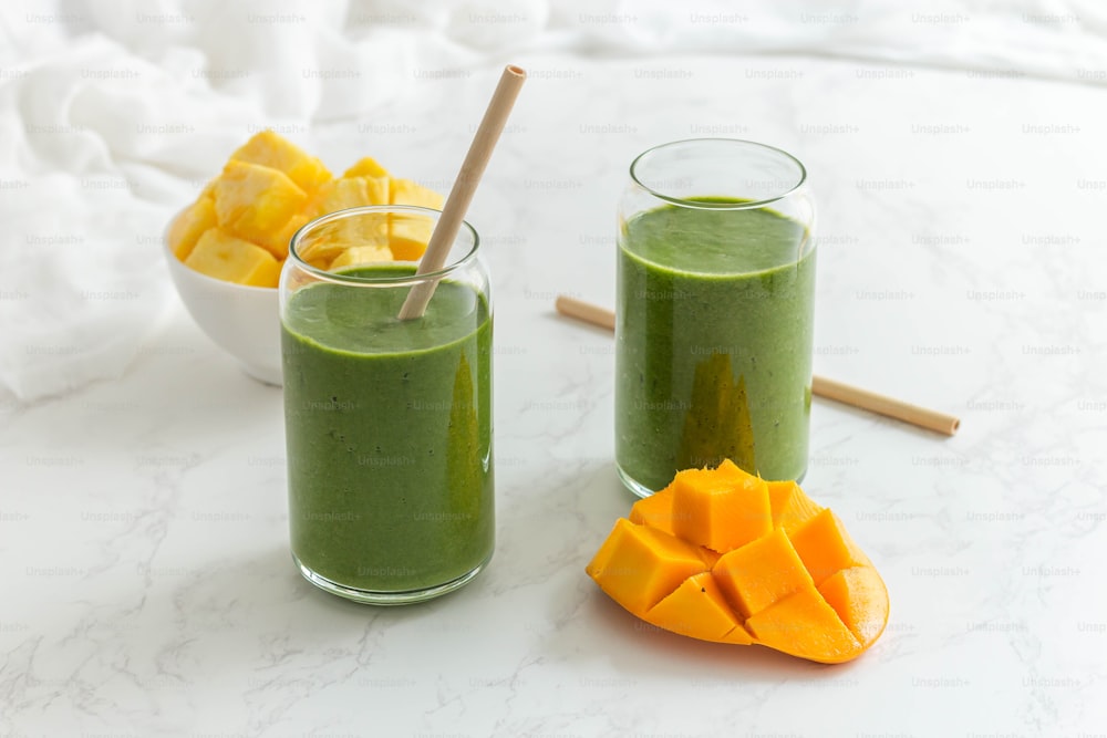 two glasses of green smoothie next to a bowl of mango slices