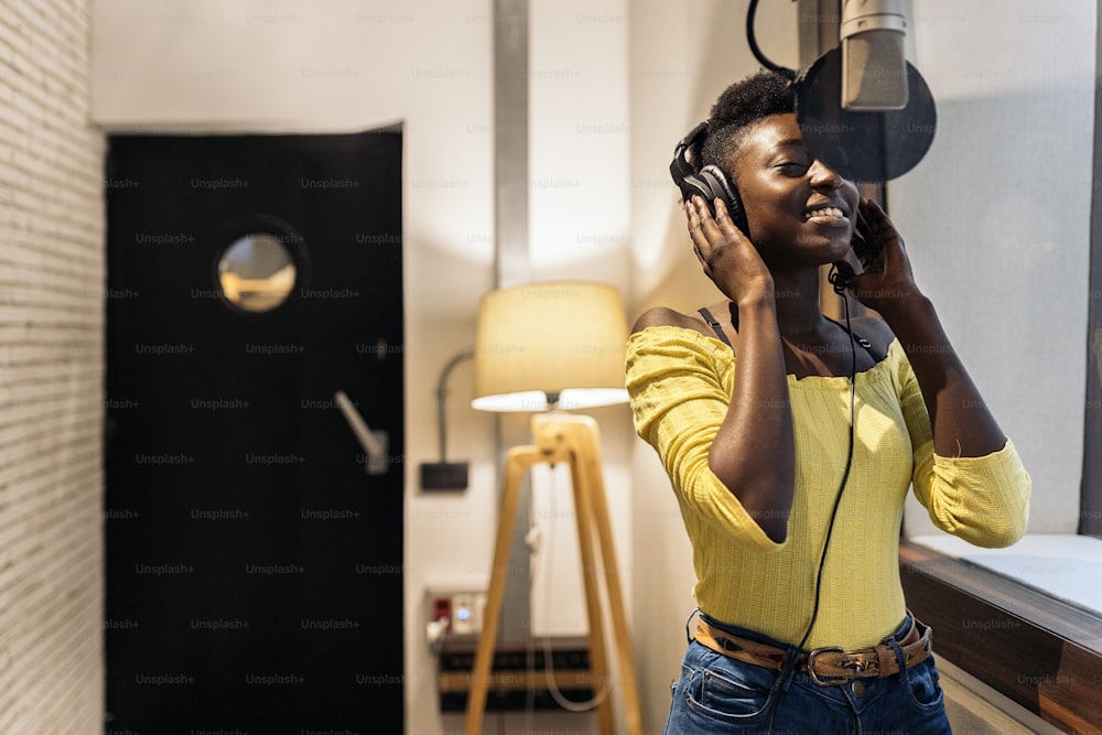 Stock photo of beautiful black woman with headphones using microphone in music studio.