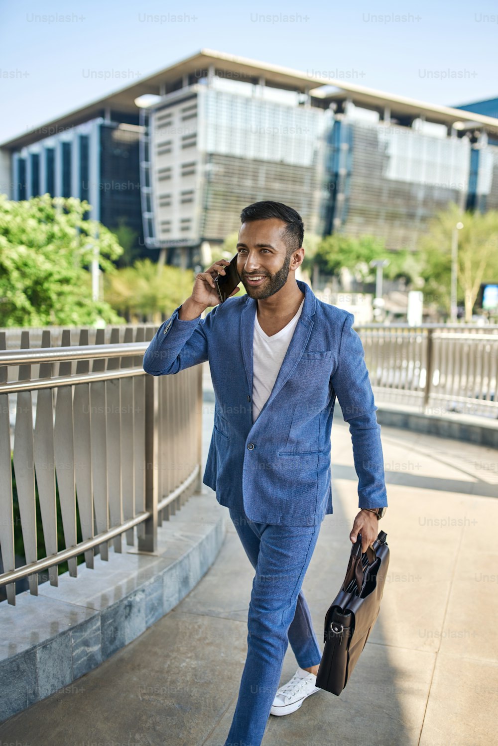 若い魅力的な笑顔のひげを生やしたインドのアラビア語のビジネスマンは、彼の朝の都市の通勤中に携帯電話を歩いて話しています