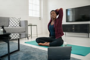 Giovane donna africana nera seduta sul tappetino da yoga nel moderno salotto di casa, allungando il collo, la spalla e la parte superiore della schiena.