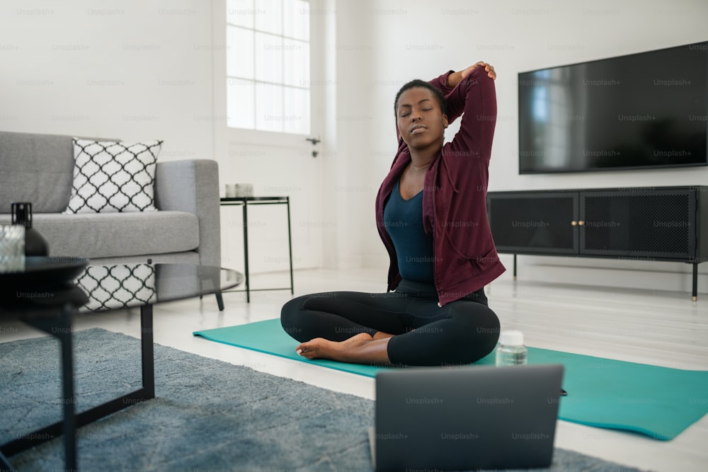 Giovane donna africana nera seduta sul tappetino da yoga nel moderno salotto di casa, allungando il collo, la spalla e la parte superiore della schiena.