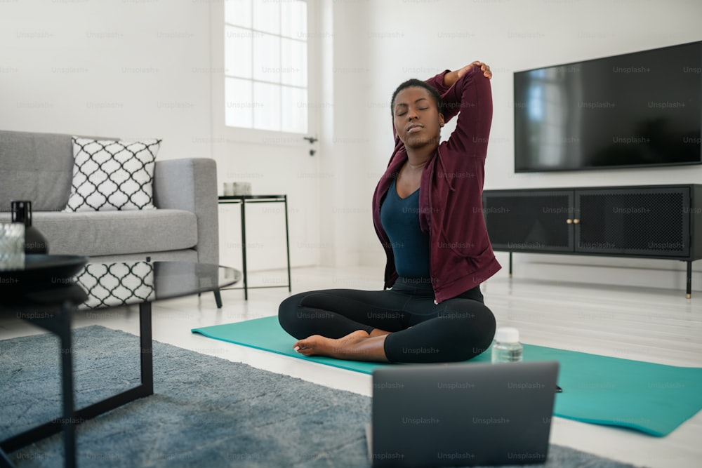 Young Black African woman sitting on yoga mat in modern lounge at home, stretching neck, shoulder and upper back.