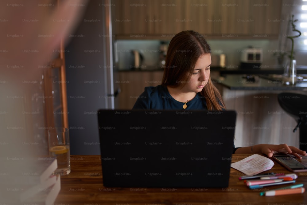 Portrait of beautiful young teenage school girl sitting at dining table, doing school work and attending virtual elearning classroom studying and learning at home