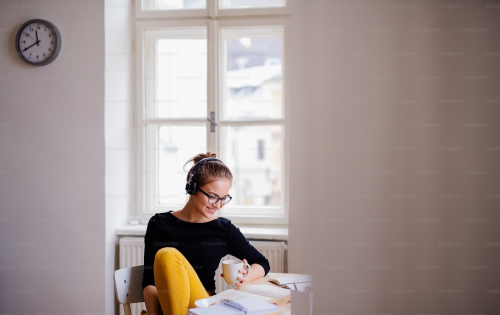 Una giovane studentessa universitaria felice seduta al tavolo di casa, che usa le cuffie durante lo studio.