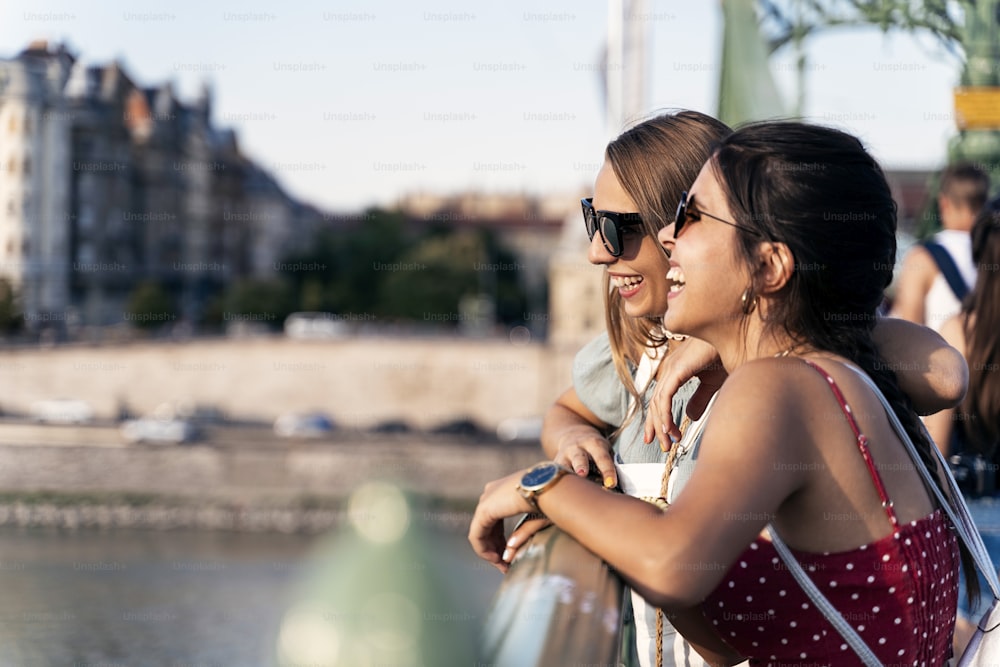 Side view of young women in sunglasses leaning on bridge railing and having good time together during walk in city