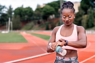 Stock photo of an African-American sprinter standing on an athletics track looking at her watch with a bottle of water and her hair tied up