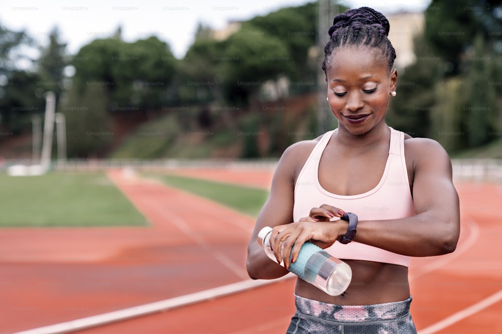 Stockfoto einer afroamerikanischen Sprinterin, die auf einer Leichtathletikbahn steht und mit einer Flasche Wasser und zusammengebundenen Haaren auf ihre Uhr schaut