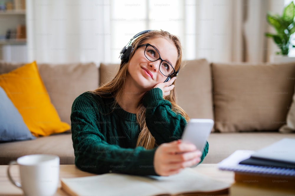 Eine junge, glückliche Studentin, die zu Hause am Tisch sitzt und beim Lernen Kopfhörer benutzt.
