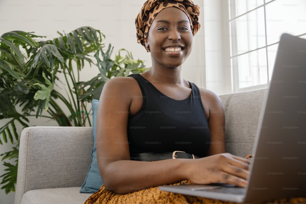 Close up Beautiful traditional young gen z African woman sitting on sofa at home, typing on laptop, smiling, looking at camera