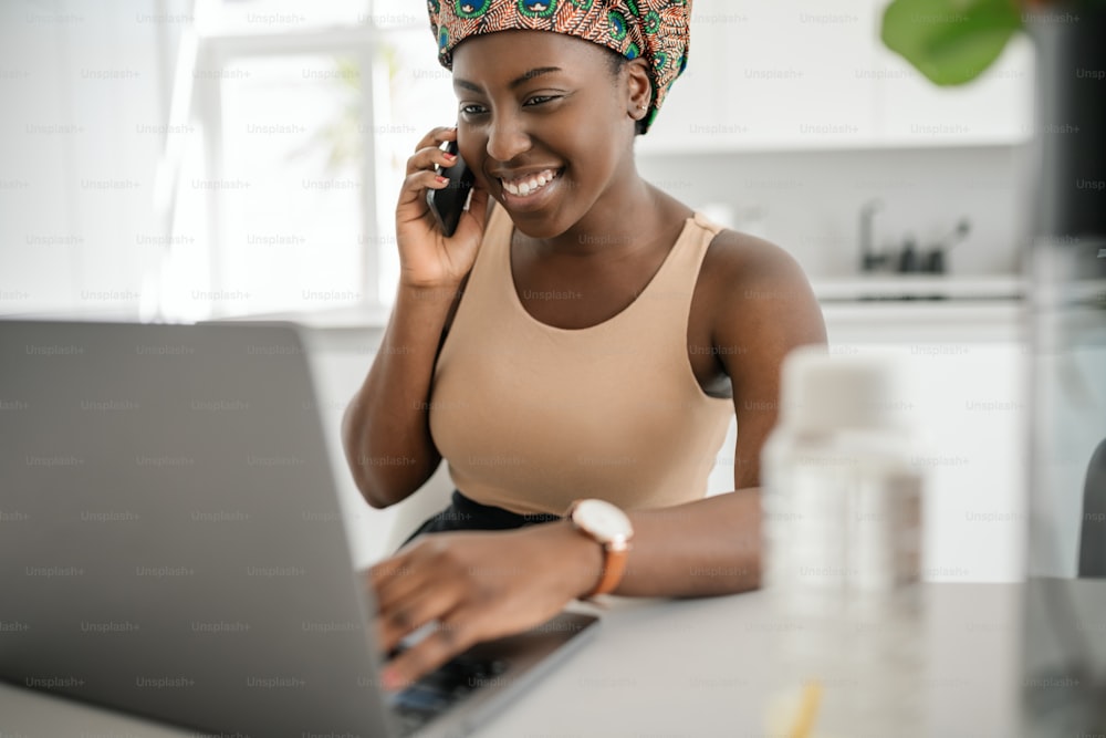 Beautiful young African woman wearing tradition headscarf. Sitting at home working on laptop. On phone call smiling and gesturing with hand