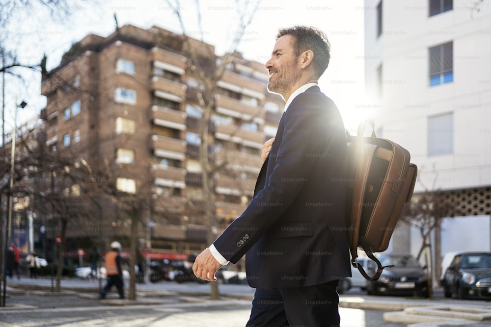 Side view of cheerful male entrepreneur in suit and with backpack walking along street on sunny day