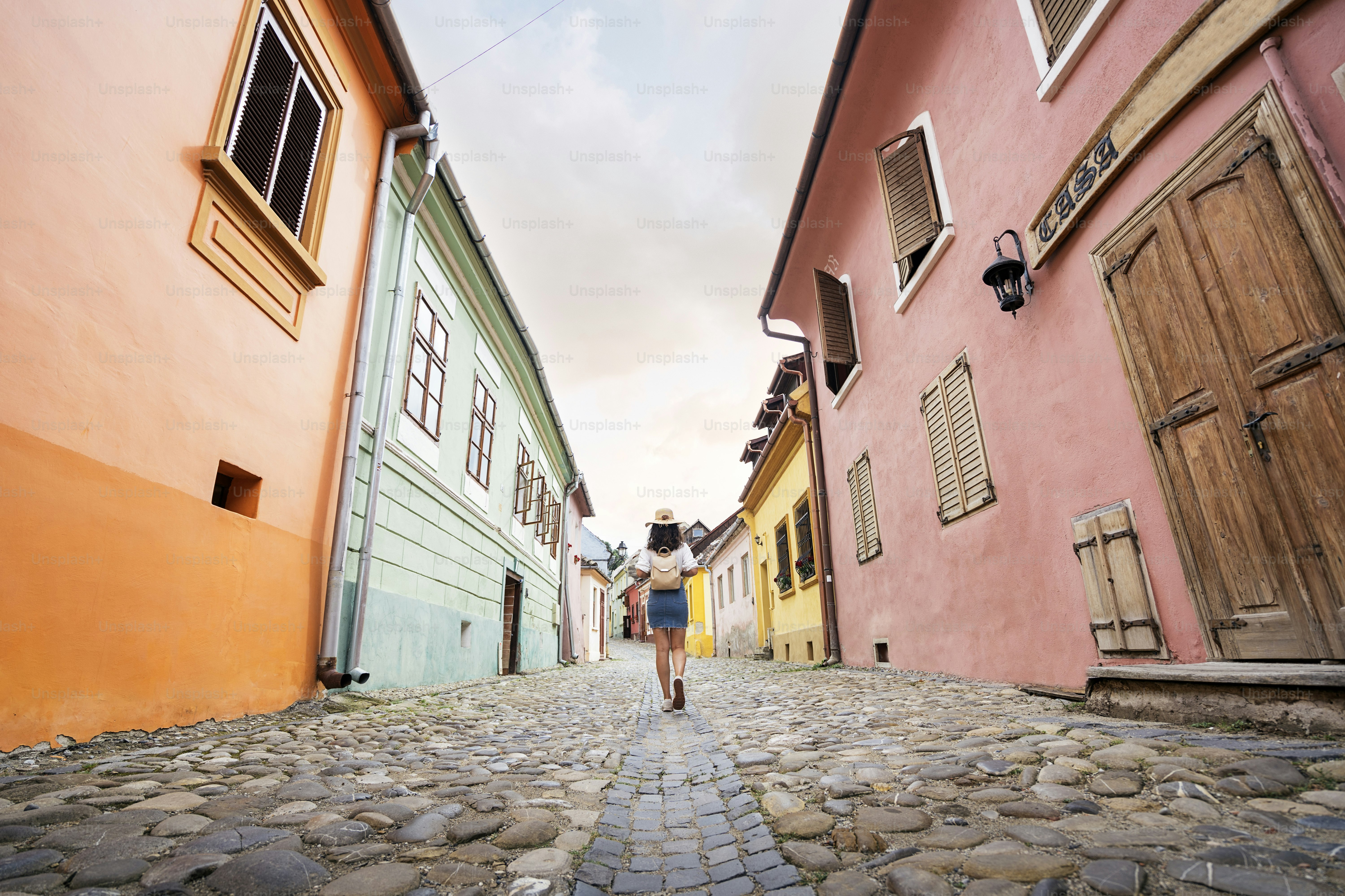 帽子をかぶった女性が、日光に照らされた明るい色とりどりの建物が並ぶ石畳の通りを歩いている後ろから
