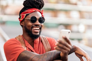 Foto stock di attraente ragazzo afroamericano con dreadlocks e una bandana usando il suo telefono. Sta ridendo.