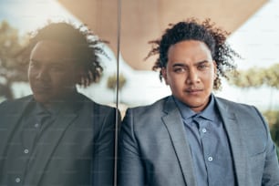 Young mixed race guy with curly afro looking seriously into camera leaning against glass wall