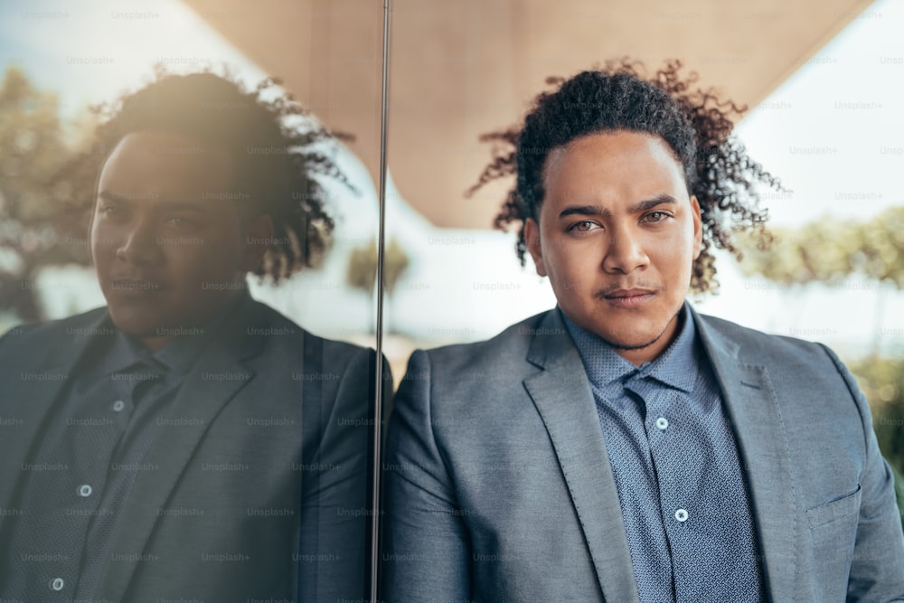Young mixed race guy with curly afro looking seriously into camera leaning against glass wall