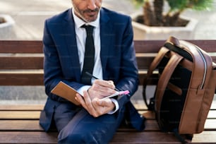 From above of mature male entrepreneur in suit writing in notebook while sitting on bench in street