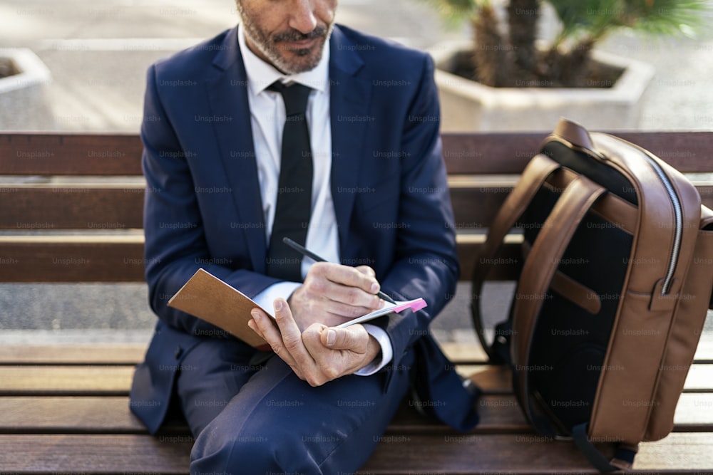 From above of mature male entrepreneur in suit writing in notebook while sitting on bench in street