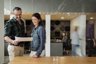 Dos empresarios sonrientes revisando el papeleo juntos mientras están de pie en un escritorio en una oficina moderna y concurrida