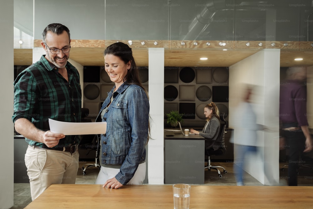 Two smiling businesspeople going over paperwork together while standing at a desk in a busy modern office