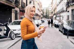 Hübsche junge muslimische Frau mit Kopftuch, die ihr Handy benutzt und durch die Stadt läuft.
