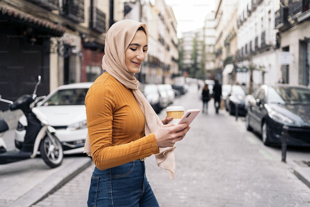 Bella giovane donna musulmana che indossa il velo usando il suo telefono cellulare e camminando per la città.