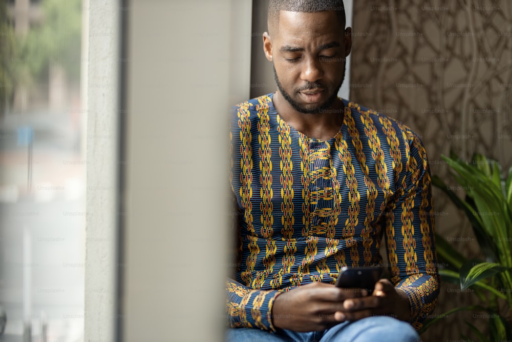Portrait of black African business man and entrepreneur sitting on window sill looking looking down and typing on mobile smart phone with copy space