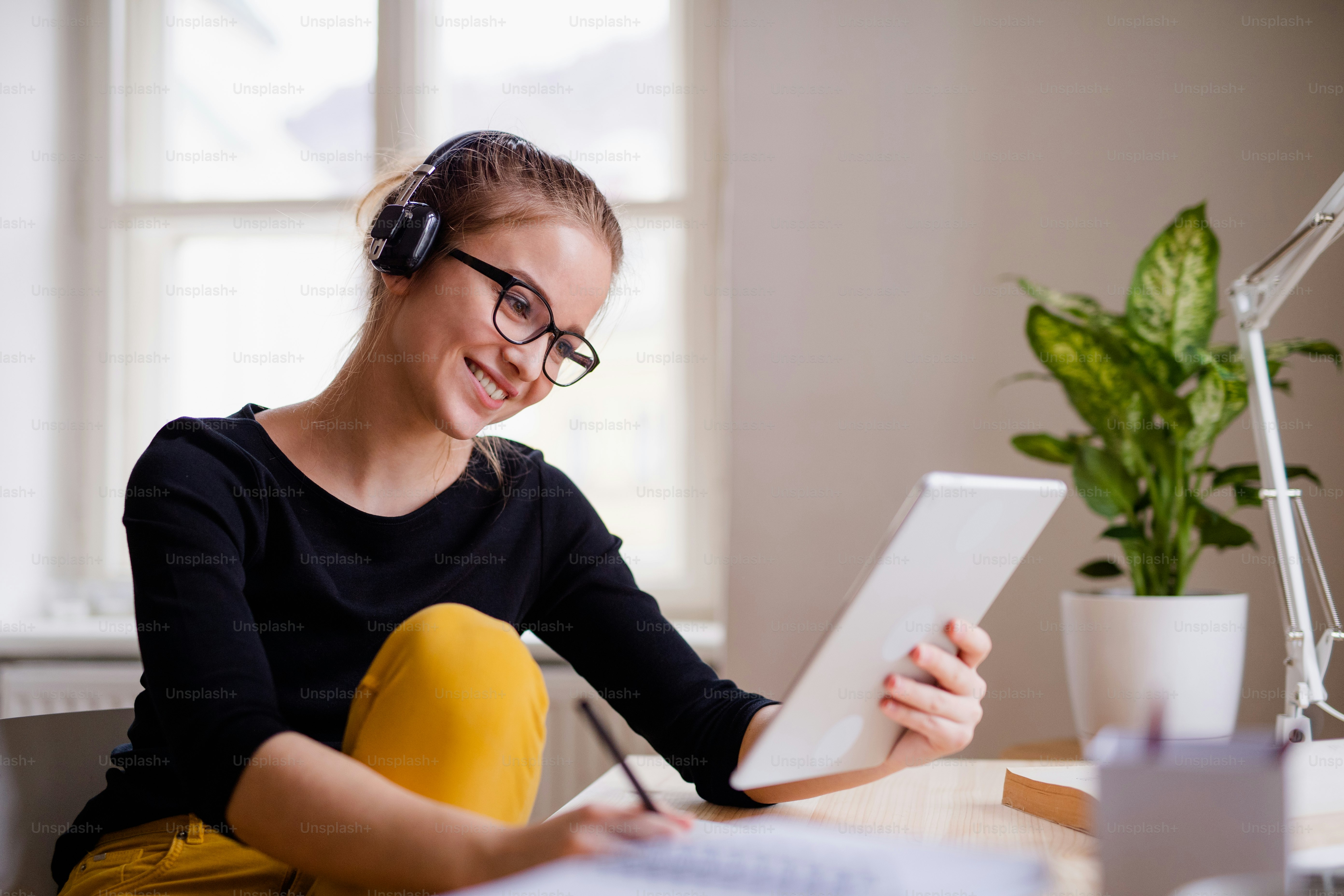 Eine junge, glückliche Studentin, die zu Hause am Tisch sitzt und beim Lernen Kopfhörer und Tablet benutzt.