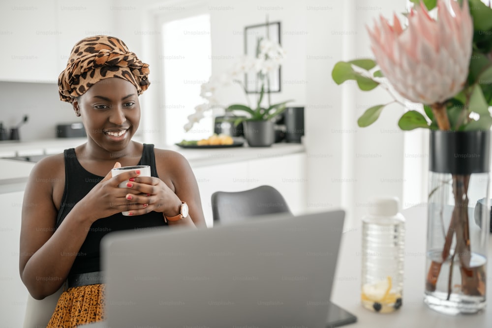 Young Black African woman at home, sitting watching her online series on laptop, holding mug of coffee