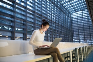 Um retrato de uma jovem estudante ou empresária sentada na mesa da sala em uma biblioteca ou escritório, usando laptop.