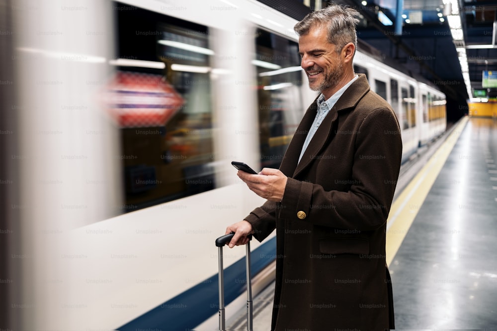Foto de archivo de una vista lateral de un hombre de negocios escribiendo en su teléfono inteligente esperando para tomar el metro en la plataforma. Está sonriendo y viste ropa informal.