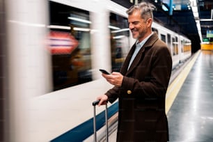 Stock photo of a side view of a business man typing in his smartphone waiting to take the underground in the platform. He is smiling and wearing casual clothes.
