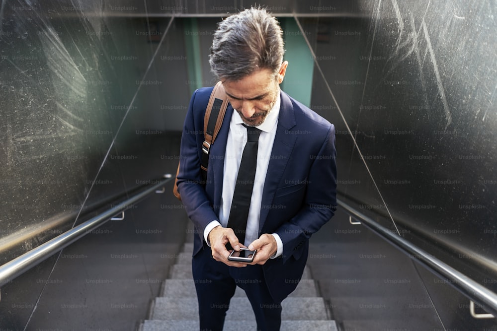 Hoher Winkel des selbstbewussten reifen männlichen Unternehmers im Anzug und mit Handy auf der Treppe der Unterführung stehend und konzentriert