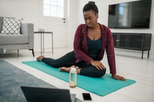 Black African woman watching online work out on laptop at home, stretching on yoga mat in living room