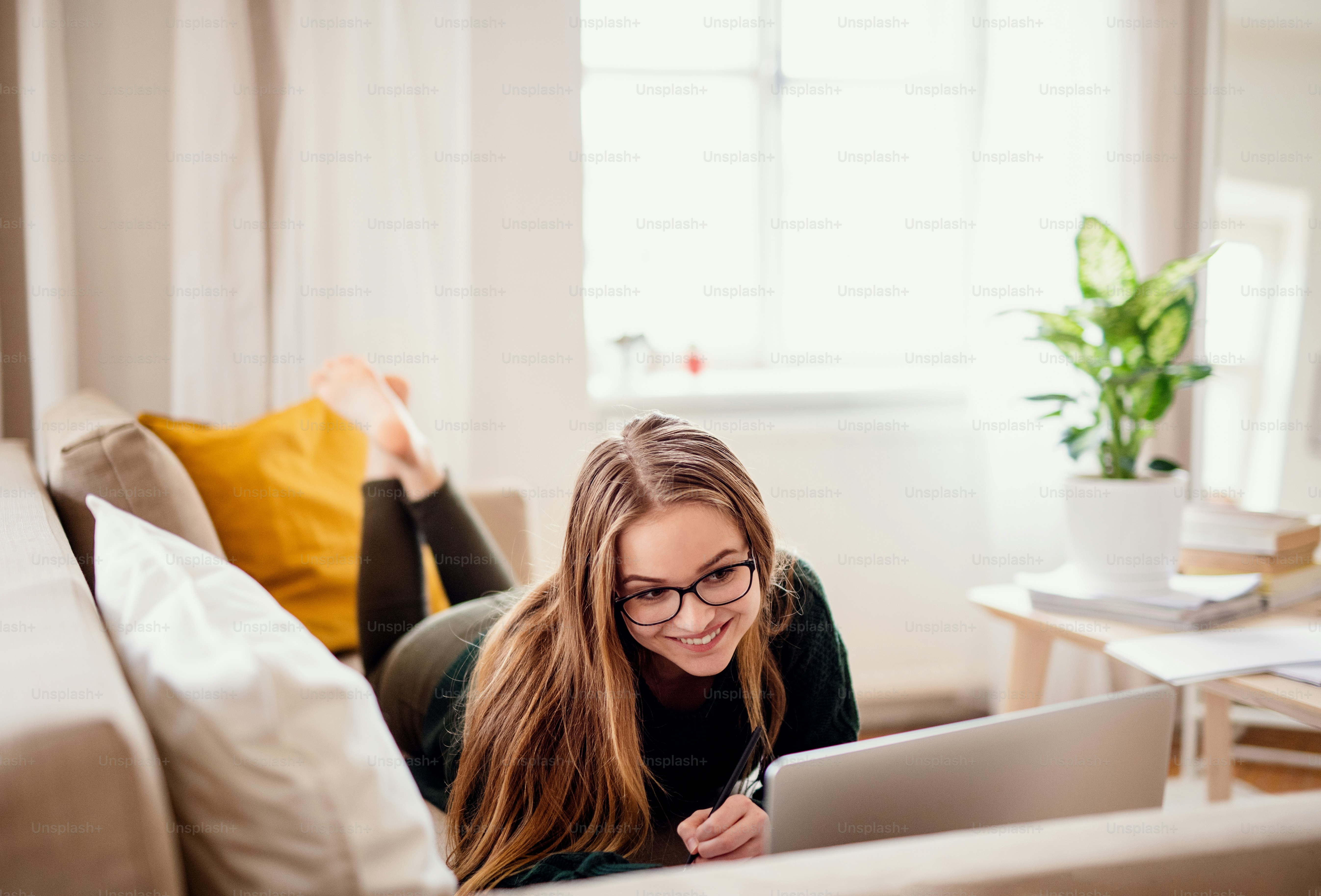Eine junge, glückliche Studentin, die zu Hause auf dem Sofa liegt und beim Lernen einen Laptop benutzt.