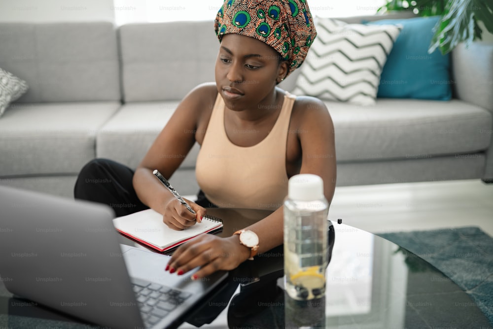 African woman studying from home, looking at laptop, writing down notes. Laptop and smartphone on home office table