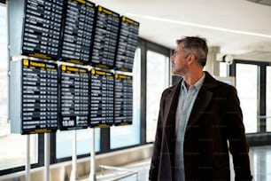 Foto de banco de imagens de um homem de negócios caucasiano olhando para o horário de partidas no aeroporto. Ele tem uma mala. Ele está usando roupas casuais.