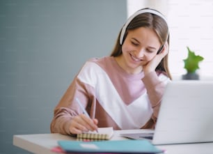 Giovane studentessa universitaria felice seduta al tavolo di casa, usando laptop e cuffie durante lo studio.