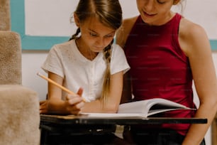 a woman and a girl sitting at a table with a book and pencil