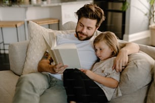 a man sitting on a couch reading a book to a little girl