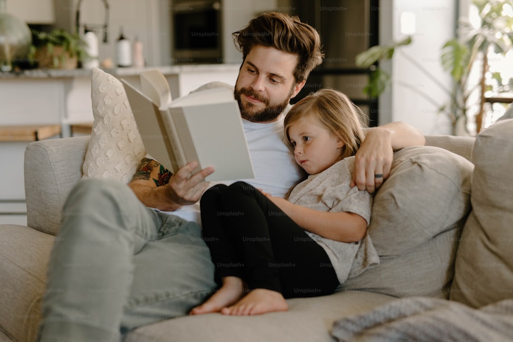 a man sitting on a couch reading a book to a little girl