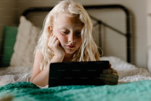 a little girl laying on a bed looking at a tablet