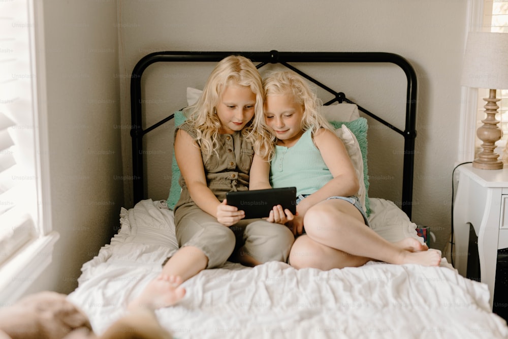 two young girls sitting on a bed looking at a tablet
