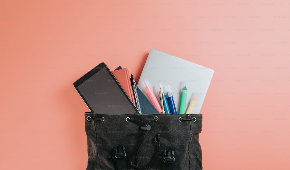 un sac noir rempli de fournitures scolaires sur fond rose