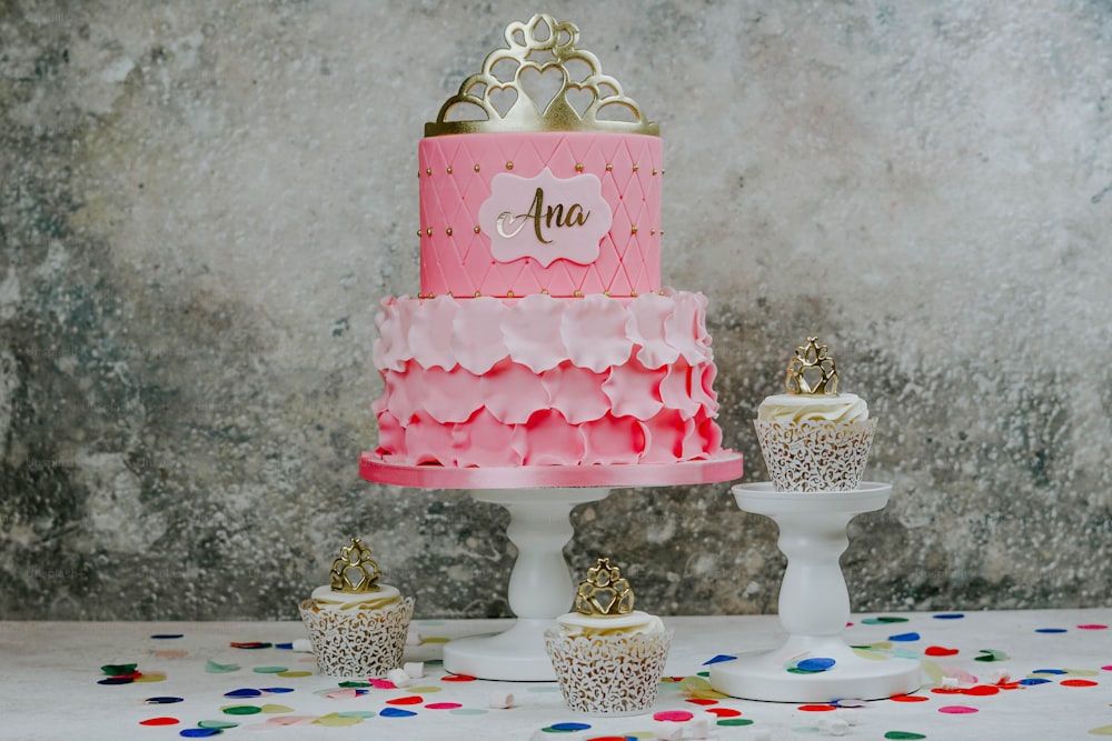a pink and gold birthday cake with cupcakes and confetti