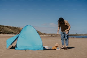 a man standing next to a blue tent on top of a sandy beach