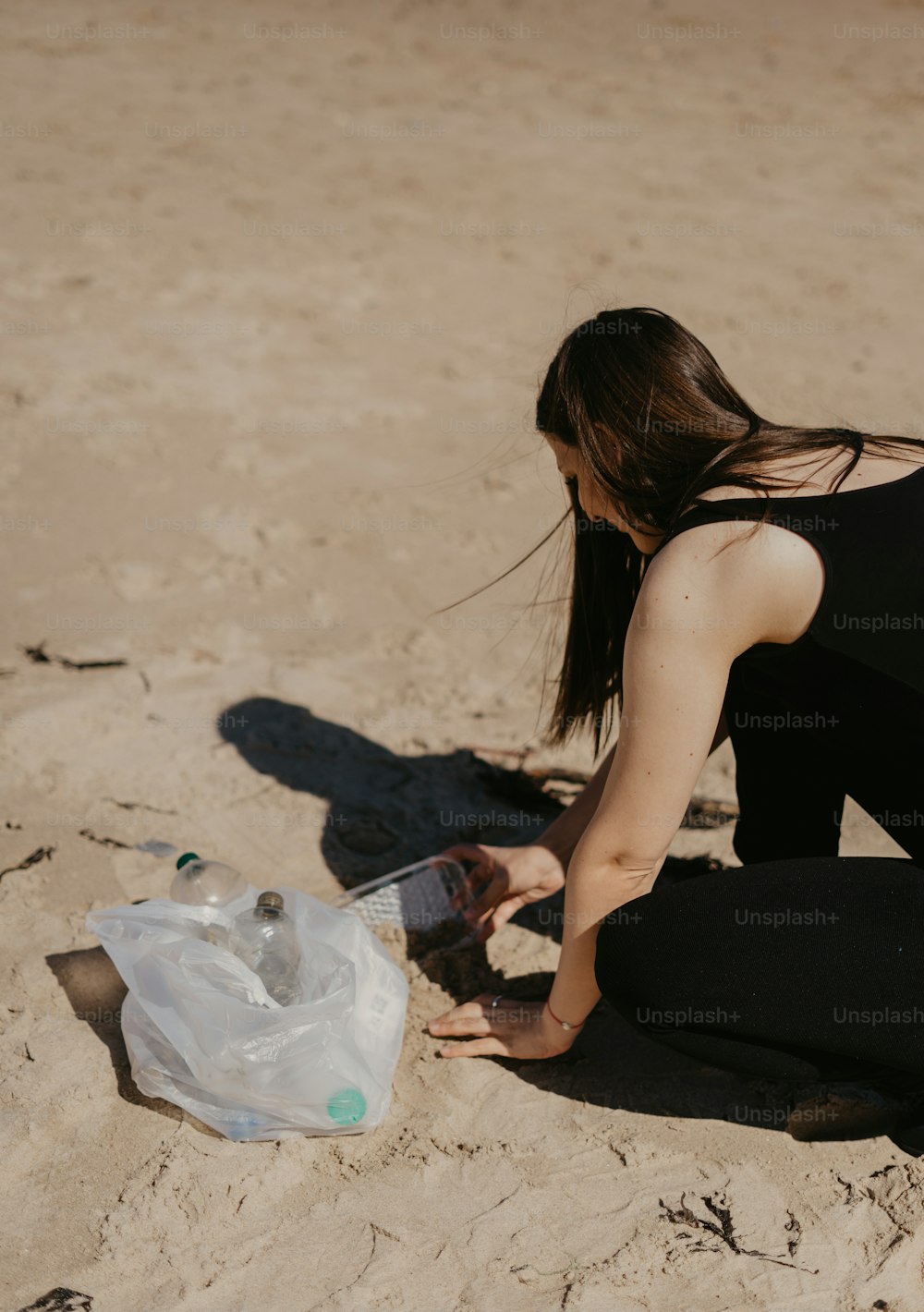 a woman kneeling down on a beach next to a plastic bottle